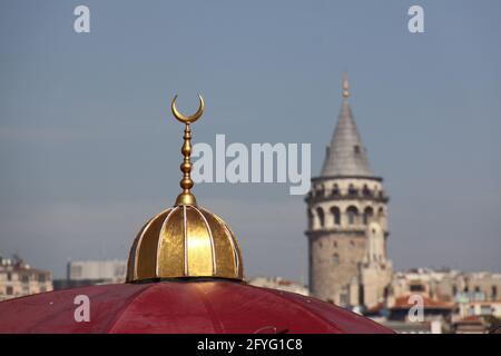 top of a red mosque dome with a golden half moon on the top and the galata tower in the background Stock Photo