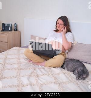 Pregnant woman on home bed with laptop talking on mobile phone Stock Photo