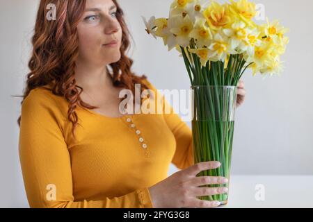 Young lady puts spring flowers in vase on white office table in room. Housewife taking care of coziness and decor. Curly hair hairstyle. Stock Photo