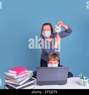 Mother of a schoolboy in a medical face mask shows on time, a school desk on a blue background during the coronavirus pandemic Stock Photo