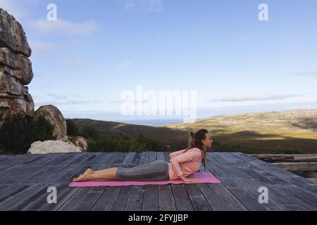 Happy caucasian woman practicing yoga lying on deck stretching in rural mountain setting