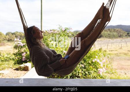 Smiling caucasian woman lying in hammock on sunny cottage terrace, looking to garden Stock Photo