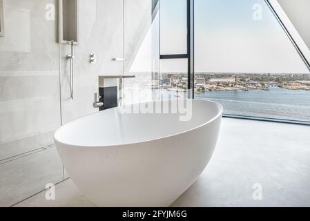 Big white soaking bathtub in spacious bathroom with shower cabin and wall window viewing city Stock Photo