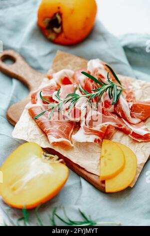 Slices of fresh prosciutto and persimmon fruit on a wooden chopping board Stock Photo