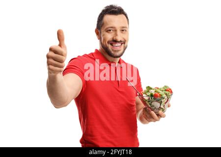 Smiling young man in a red t-shirt eating a healthy fresh salad and showing thumbs up isolated on white background Stock Photo