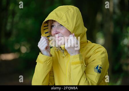 Woman in a yellow raincoat with the hood p looking into the distance sat on a bench in woodland. Stock Photo