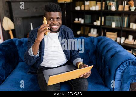 Contented bearded Afro American male freelancer relaxing on sofa smiling during the phone call after online meeting with colleagues.Businessman Stock Photo
