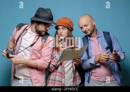 Tourists, two men and one woman, perplexedly look at the map and plan a route for a walk and sightseeing. Blue background. Stock Photo