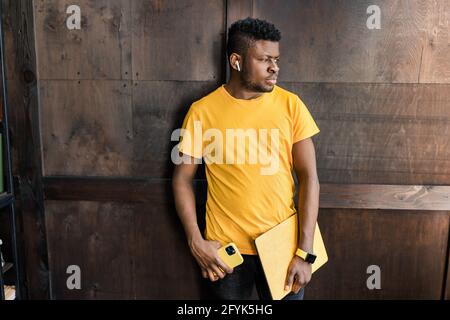 Serious focused afro american freelancer is standing near the wall, with yellow laptop, smart phone in hands, wearing casual trendy yellow and black Stock Photo