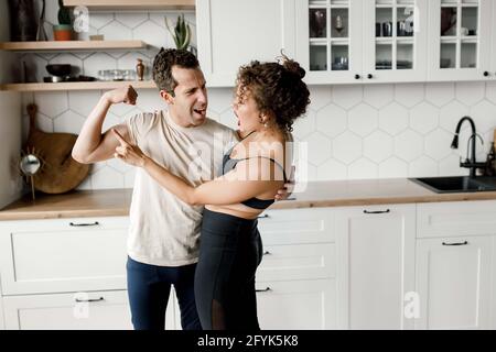 Joyful young sport couple in activewear looking at each other while hugging, laughing showing biceps and strength after training at home. Healthy Stock Photo