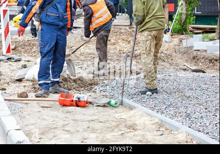 Road workers use shovels to prepare a sandy foundation on a sidewalk and cover it with rubble along concrete curbs on a clear day. Copy space. Stock Photo