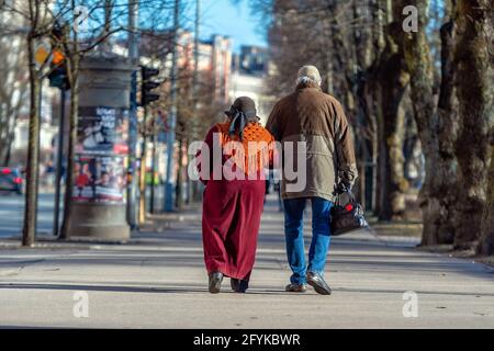 A couple of eldery people in hands are walking through the city street. Rear view. Stock Photo