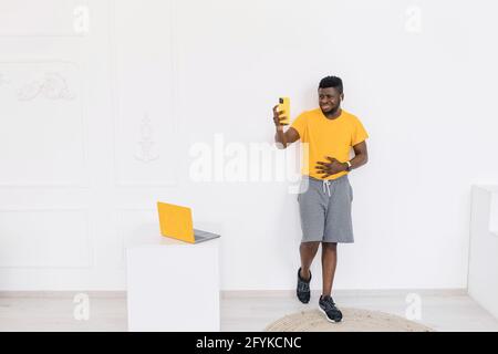 Smiling Afro American student or freelancer standing near the wall in trendy yellow t-shirt, having online call with smart phone with friends or Stock Photo