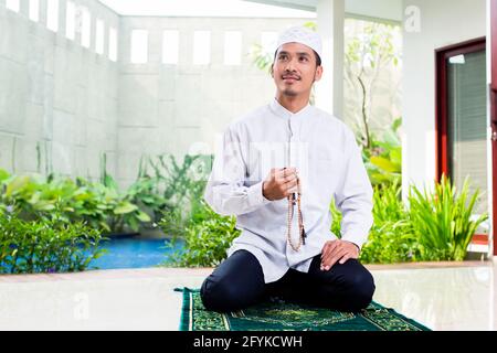 Asian Muslim man praying at home sitting on prayer carpet in his house in front of the tropical garden Stock Photo