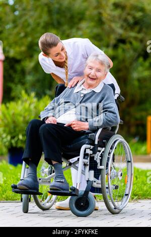 Nurse pushing senior woman in wheelchair on walk thru garden in summer Stock Photo