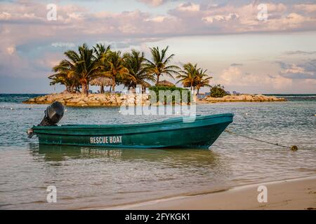 Small dark green rescue boat with motor tied at shore in front of very small tropical island with palm trees, small beech with plank beds for sunbathi Stock Photo