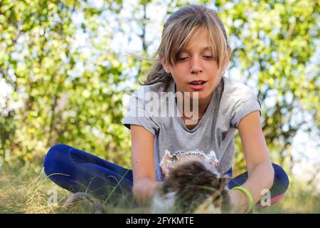Defocus blonde little expression girl playing and caress cat, black and white small kitten. Nature blurred green summer background. Girl playing pet. Stock Photo