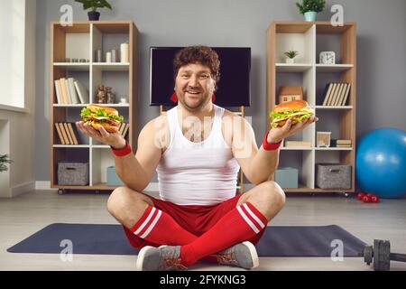 Happy chubby man sitting on sports mat, eating fast food and enjoying his cheat day Stock Photo