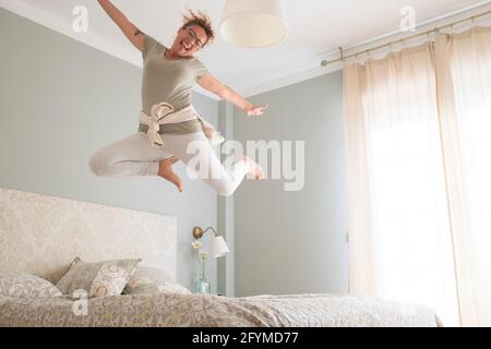Young adult pretty woman go crazy for happiness and jump on the bed with coffee cup - overjoyed female caucasian people alone at home have fun jumping Stock Photo