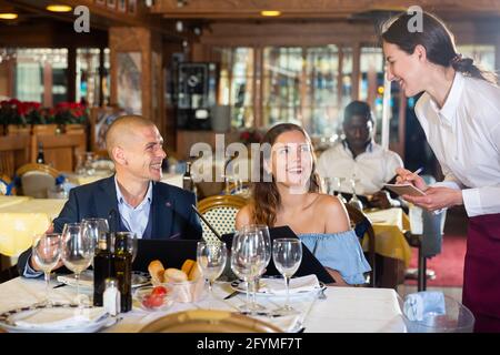 Young positive waitress taking table order from guests, helping couple in choosing meals from menu Stock Photo