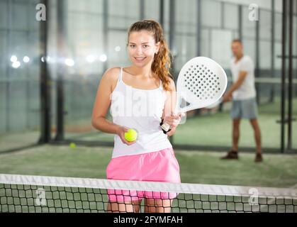 Portrait of smiling sporty girl with paddle tennis racquet and ball in her hands ready to play match standing on indoor court on blurred background of Stock Photo