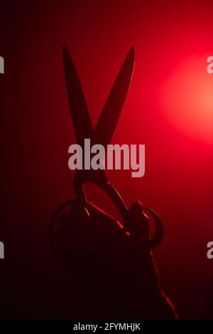 Vertical shot of a hand holding a large pair of scissors with a bright red run shining behind Stock Photo