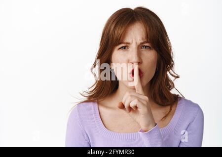 Close up portrait of angry shushing girl, say shhh and frowning, press finger to lips, taboo dont speak gesture, telling to be quiet, standing against Stock Photo