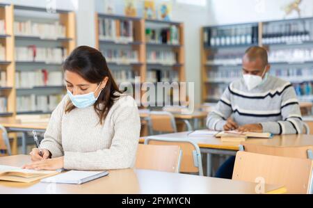Interested young Latina in medical face mask spending time in library during coronavirus pandemic, reading books and making notes. Compelled precautio Stock Photo