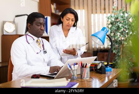 Focused african american doctor working in clinic office with female assistant, filling out medical forms on laptop Stock Photo