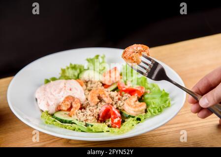 Salad with shrimps, quinoa, tomatoes, peppers, cucumber, lettuce, mayonnaise on white round plate on wooden table. Stock Photo