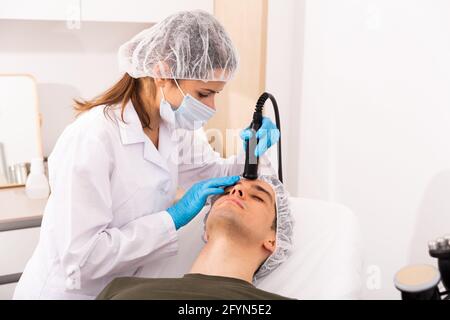 Young man receiving face rejuvenation treatment on modern equipment at cosmetology clinic, male skincare concept Stock Photo