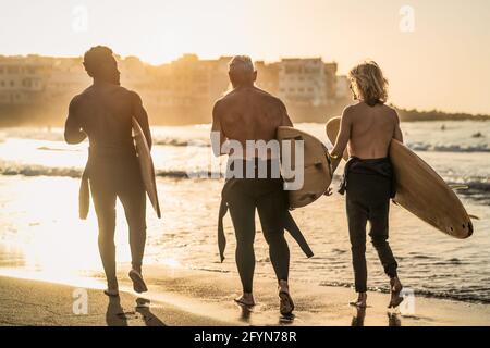 Happy fit surfers with different age and race having fun during surf day on the beach at sunset time - Extreme sport lifestyle and friendship concept Stock Photo
