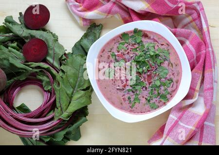 Beetroot soup in white bowl decorated green parsley next to fresh raw beetroots on wooden table top view Stock Photo