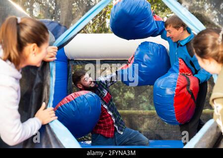 Friends having fun in outdoor amusement park, women watching men in big boxing gloves boxing on inflatable ring Stock Photo