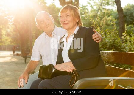 Happy senior man and woman sitting hugging on bench in green park Stock Photo
