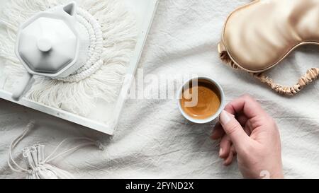 Espresso in hand, tray with white ceramic espresso coffee maker on trendy macrame pad. Sleep mask on crumpled bed sheets. Female hand with cup of Stock Photo
