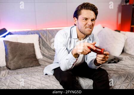 Concentrated guy playing video game, holding remote red controller, enjoying leisure time with device, engaged in online competition with friends Stock Photo