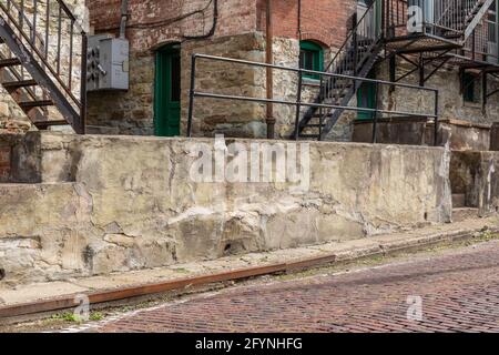 Crumbling concrete wall alongside a red brick street, brick apartment building behind, horizontal aspect Stock Photo