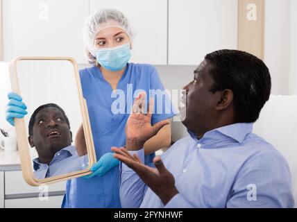 Indignant African American looking at mirror examining his face after cosmetology procedures on background with confused young female doctor Stock Photo