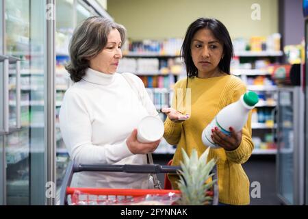 Interested Hispanic woman shopping in supermarket, choosing dairy products, asking for advice from older woman Stock Photo