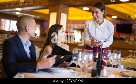 Attractive smiling woman waiter receiving order from guest in fashionable restaurant Stock Photo
