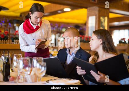 Attractive smiling woman waiter receiving order from guest in fashionable restaurant Stock Photo