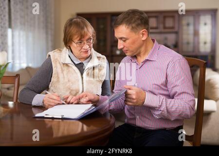 Adult man with his elderly mother discussing documents while sitting at home table Stock Photo