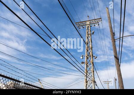 Multiple metal and wood power poles with lights and cables by a chain link fence against a blue sky with clouds, horizontal aspect Stock Photo