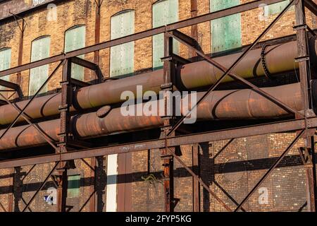 Brightly lit facade of a warehouse building with large pipes running along the exterior, green fiberglas window covers, rusting industrial landscape, Stock Photo