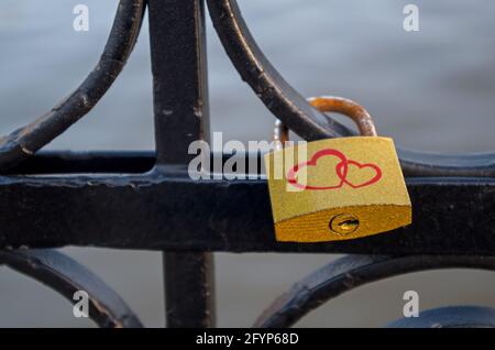 Golden padlock with two red hearts and rusty bracket hangs on black rounded swirls of bridge fence across river on a blurred background of water. Conc Stock Photo