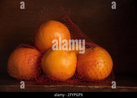 Oranges in red netting are displayed on a wooden shelf in a still life image.