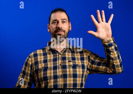 Handsome bearded man in a yellow plaid shirt waving with one hand isolated on blue studio background. Kindness concept Stock Photo