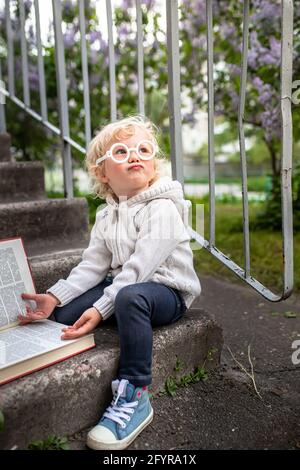 little girl reads a big book. encyclopedia lies on steps of the school. Getting knowledge by children in kindergarten. Kid looks up