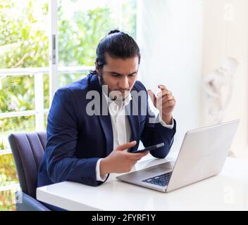 young latino Man with laptop texting on mobile phone sitting at table. Home working Stock Photo